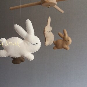 May include: A white and brown felt bunny mobile with four bunnies hanging from a wooden arm. The bunnies have closed eyes and are in a flying position.