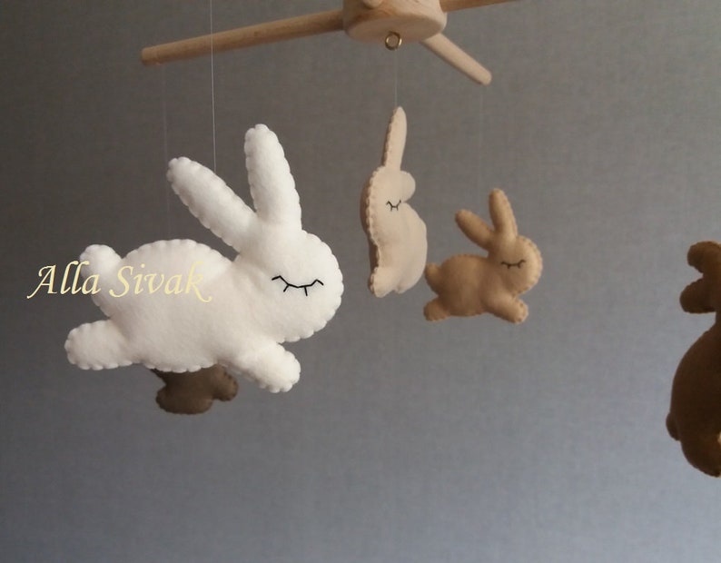 May include: A white and brown felt bunny mobile with four bunnies hanging from a wooden arm. The bunnies have closed eyes and are in a flying position.