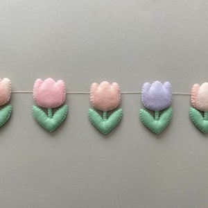 May include: A garland of felt tulips in pink, light pink, lavender, and green. The tulips are strung together with white thread.