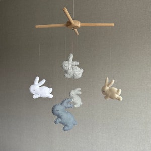 May include: A wooden baby mobile with five felt bunny toys. The bunnies are in shades of gray, white, and cream. The mobile is hanging from a wooden frame.