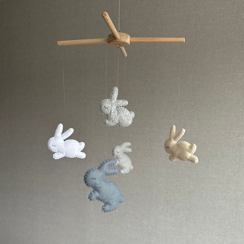 May include: A wooden baby mobile with five felt bunny toys. The bunnies are in shades of gray, white, and cream. The mobile is hanging from a wooden frame.