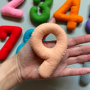 May include: A set of felt numbers in various colours, including pink, green, orange, red, blue, and peach. The numbers are arranged in a semi-circle shape, with the number nine in the centre.