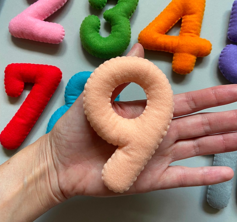 May include: A set of felt numbers in various colours, including pink, green, orange, red, blue, and peach. The numbers are arranged in a semi-circle shape, with the number nine in the centre.