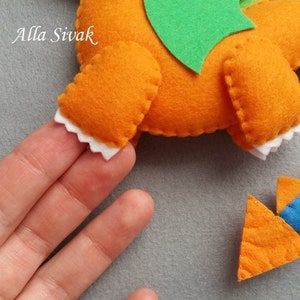 May include: A felt orange dragon with green wings and white teeth. The dragon is held in a hand. The dragon's tail is a separate piece and is orange with blue spikes.