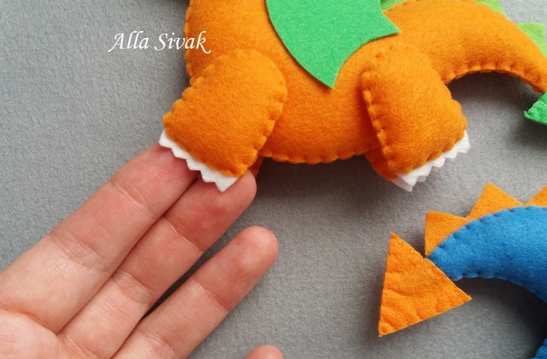 May include: A felt orange dragon with green wings and white teeth. The dragon is held in a hand. The dragon's tail is a separate piece and is orange with blue spikes.