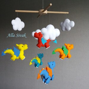 May include: A baby mobile with five felt dragons and two white clouds. The dragons are yellow, orange, red, and blue. The mobile is suspended from a wooden dowel rod.