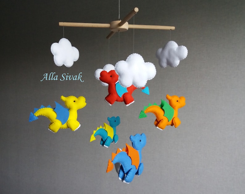 May include: A baby mobile with five felt dragons and two white clouds. The dragons are yellow, orange, red, and blue. The mobile is suspended from a wooden dowel rod.