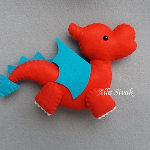 May include: Red felt dragon toy with blue felt wings and tail. The dragon has a black button eye and white felt teeth.