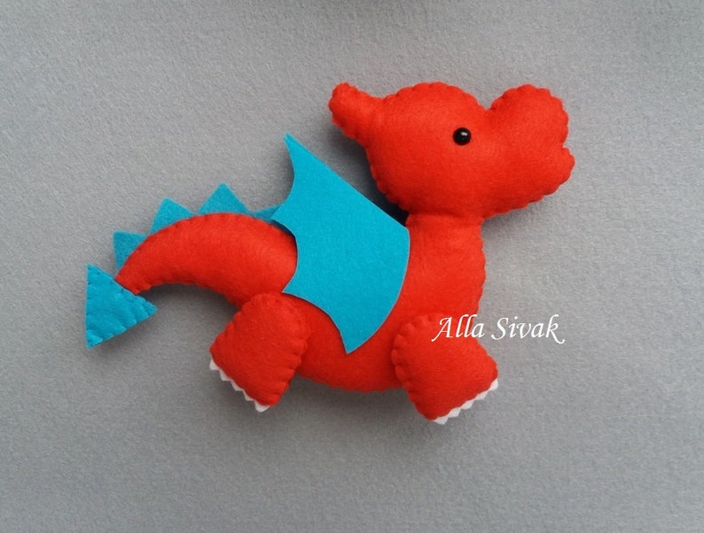 May include: Red felt dragon toy with blue felt wings and tail. The dragon has a black button eye and white felt teeth.