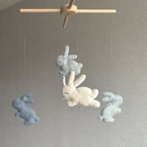 May include: A wooden baby mobile with four felt bunny toys. The bunnies are in shades of gray and white. The mobile is hanging from a wooden crossbar.