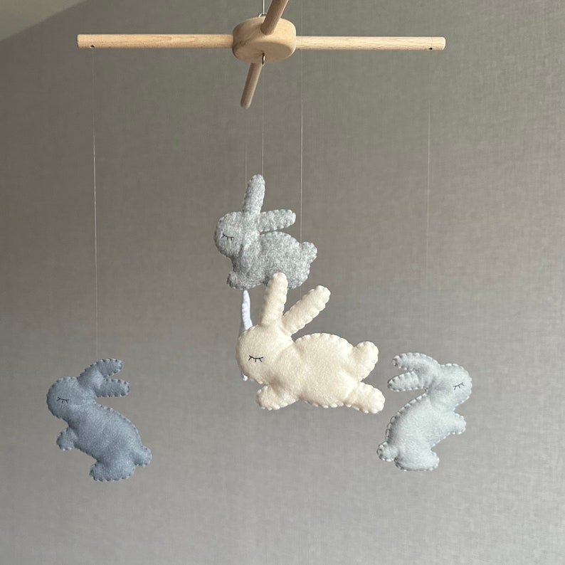 May include: A wooden baby mobile with four felt bunny toys. The bunnies are in shades of gray and white. The mobile is hanging from a wooden crossbar.