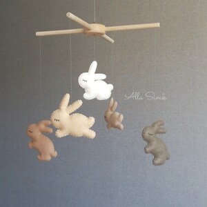 May include: A wooden mobile with five felt bunny toys hanging from it. The bunnies are in shades of brown and white. The mobile is suspended from a wooden crossbar.