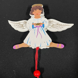 May include: A wooden angel figurine with white wings, a white dress, and a pink ribbon. The angel is holding a small, brown object in its hands. The angel is attached to a red string with a red bead.