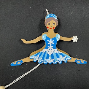 May include: A blue and white wooden ballerina doll with a snowflake design on her dress. She is wearing a crown and has brown hair. The doll is in a split position with her arms outstretched.