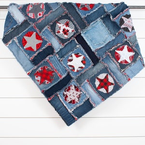 May include: A denim quilt with a patchwork design featuring red, white and black stars. The quilt is hanging on a white wooden wall.