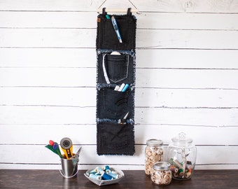 Op de afbeelding: Een denim wandorganizer met vier vakken, hangend aan een witte muur. De vakken zijn gevuld met verschillende items, waaronder pennen, potloden en spoelen garen.