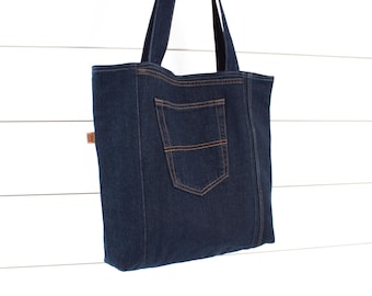 May include: A dark blue denim tote bag with a large pocket on the front. The bag has brown stitching and a brown leather tag.