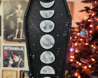 May include: Black coffin-shaped wall hanging featuring a celestial design. The artwork depicts the phases of the moon in white against a black background, with white speckles. A jute string is attached for hanging. A bat-shaped decoration is visible in the background.