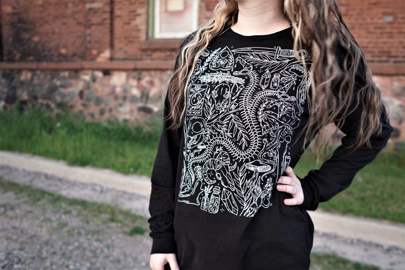 Pode incluir: Camiseta preta de manga comprida com uma estampa gr&aacute;fica branca de uma cobra esquel&eacute;tica, plantas e outros s&iacute;mbolos m&iacute;sticos. O texto "The Moon" &eacute; vis&iacute;vel na camiseta.