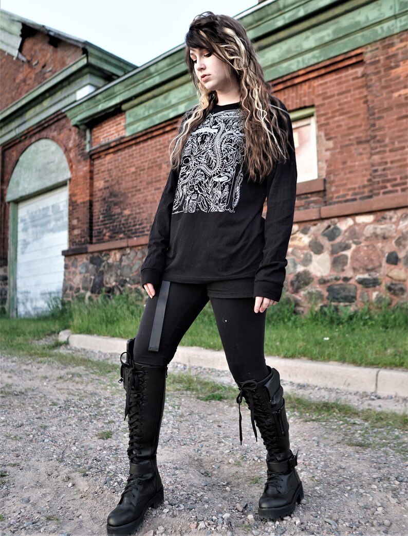 Pode incluir: Uma mulher vestindo uma camisa preta de manga comprida com uma estampa gr&aacute;fica branca, leggings pretos e botas pretas de cano alto. Ela est&aacute; em p&eacute; na frente de um pr&eacute;dio de tijolos.