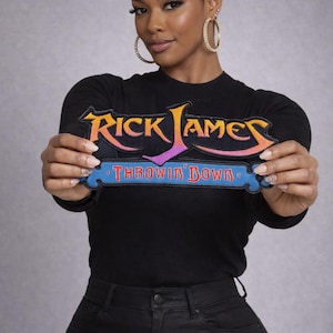 May include: Black long-sleeve shirt with a colorful "Rick James Throwin' Down" patch. The patch features orange, purple, and blue text. The person is wearing black jeans and large hoop earrings.