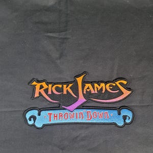 May include: Embroidered patch with the text "Rick James" in orange, yellow, and purple, above a blue banner with the words "Throwin' Down" in red. The patch is set against a black background.