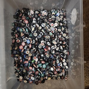 Pode incluir: Um recipiente de plástico transparente cheio de uma grande variedade de fichas de poker. As fichas são principalmente pretas com vários desenhos, números e símbolos coloridos. As fichas são redondas e empilhadas.