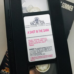 May include: A black VHS tape case with a white label. The label reads "MGM/UA Home Video" and "A Shot in the Dark". The case also includes details about the movie, such as the color, rating, runtime, and sound system. The copyright information is also included.