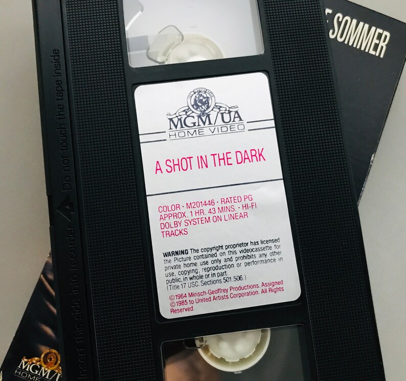 May include: A black VHS tape case with a white label. The label reads "MGM/UA Home Video" and "A Shot in the Dark". The case also includes details about the movie, such as the color, rating, runtime, and sound system. The copyright information is also included.