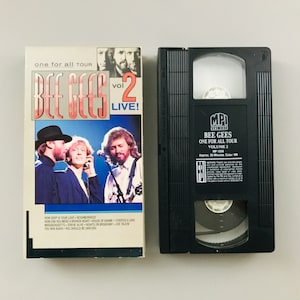 May include: A black VHS tape with a white label that reads "Bee Gees One For All Tour Volume 2" with a picture of the band on the front. The tape is approximately 50 minutes long and is in color.
