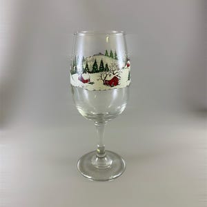 May include: A clear glass wine glass with a winter scene painted on it. The scene features a snowy landscape with houses, trees, and snow falling.