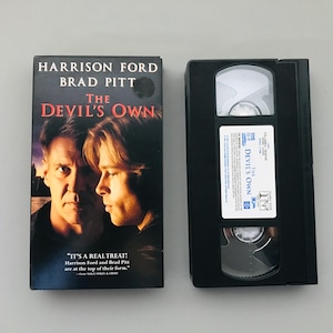 May include: VHS video cassette tape of the movie "The Devil's Own" starring Harrison Ford and Brad Pitt. The cover features a close-up of the actors' faces. The quote "It's a real treat! Harrison Ford and Brad Pitt are at the top of their form." is printed on the bottom of the cover.