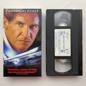 May include: A VHS tape case for the movie Air Force One starring Harrison Ford. The case is black with a white label featuring the movie title and a photo of Harrison Ford. The text "Breathtaking, gripping, nail biting, edge-of-your-seat entertainment!" is printed on the case.