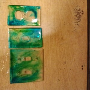 May include: Three light switch covers with a green and blue marbled resin design. The covers are made of clear resin with a swirling pattern of green and blue.