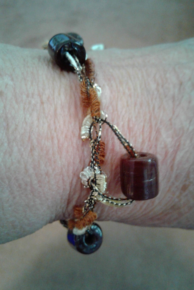May include: A brown and black beaded bracelet with gold and white accents. The bracelet is made with a variety of materials, including beads, yarn, and string.