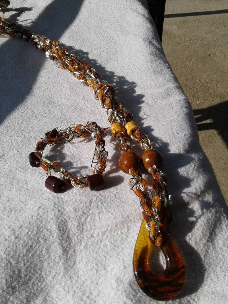 May include: A brown and gold crocheted necklace with wooden beads and a brown glass pendant. A matching brown and gold crocheted bracelet is also included.