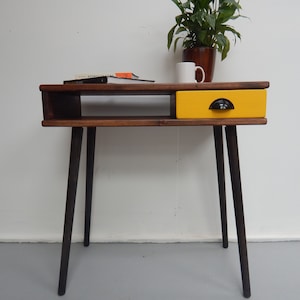 May include: A wooden desk with a yellow drawer and black legs. The desk has a shelf underneath the top surface. A plant in a brown pot and a white mug are on the desk.
