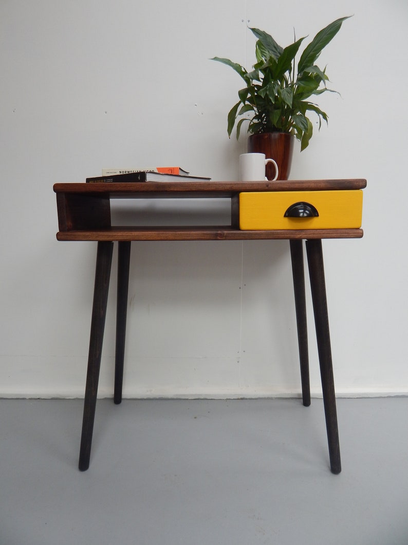 May include: A wooden desk with a yellow drawer and black legs. The desk has a shelf underneath the top surface. A plant in a brown pot and a white mug are on the desk.