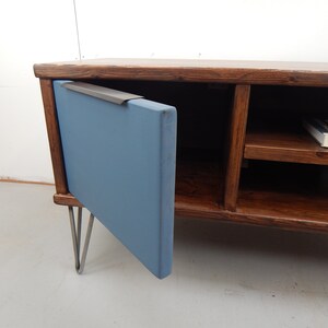 May include: A wooden TV stand with a blue door and hairpin legs. The door has a metal handle and the stand has two shelves. A book titled "NORWEGEN" is on the top shelf.
