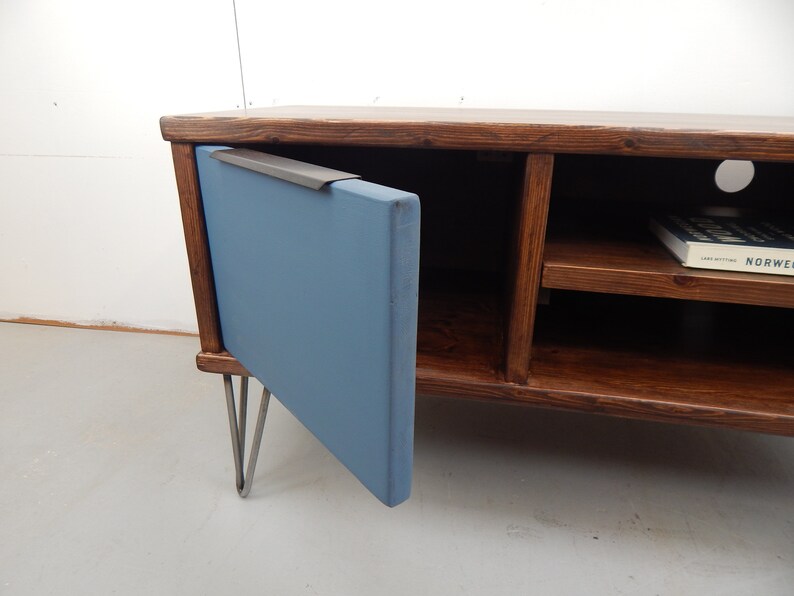 May include: A wooden TV stand with a blue door and hairpin legs. The door has a metal handle and the stand has two shelves. A book titled "NORWEGEN" is on the top shelf.