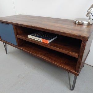 May include: A dark brown wooden TV stand with a blue door and two shelves. The stand has hairpin legs and a book titled "Norwegian Wood" on the top shelf.