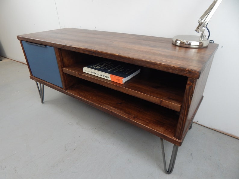 May include: A dark brown wooden TV stand with a blue door and two shelves. The stand has hairpin legs and a book titled "Norwegian Wood" on the top shelf.