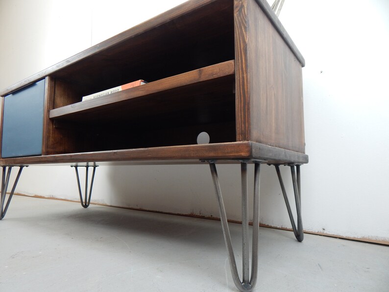 May include: A dark brown wooden TV stand with a blue door and a shelf. The stand has hairpin legs and is sitting on a light grey floor.