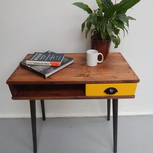May include: A wooden side table with a yellow drawer and black legs. The table has a shelf and a book titled "Norwegian Wood" is on top of the table.