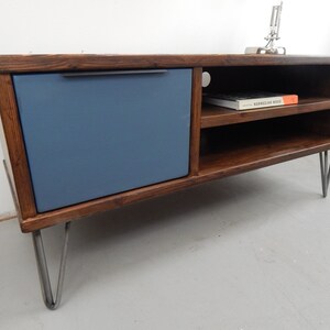 May include: A dark wood media console with a blue door and two shelves. The console has hairpin legs and a book titled "Norwegian Wood" on the top shelf.