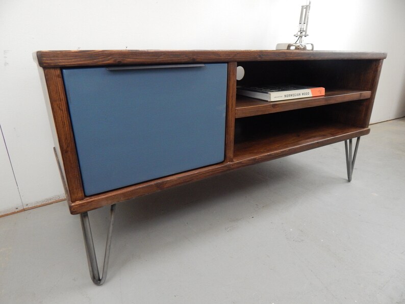 May include: A dark wood media console with a blue door and two shelves. The console has hairpin legs and a book titled "Norwegian Wood" on the top shelf.