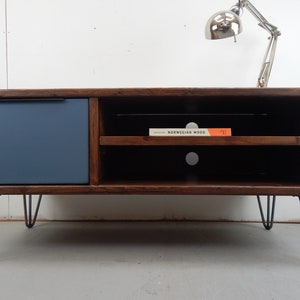 May include: A dark brown wooden TV stand with a blue painted drawer and two shelves. The stand has hairpin legs and is sitting on a light grey floor. A book titled "Norwegian Wood" is sitting on the top shelf.