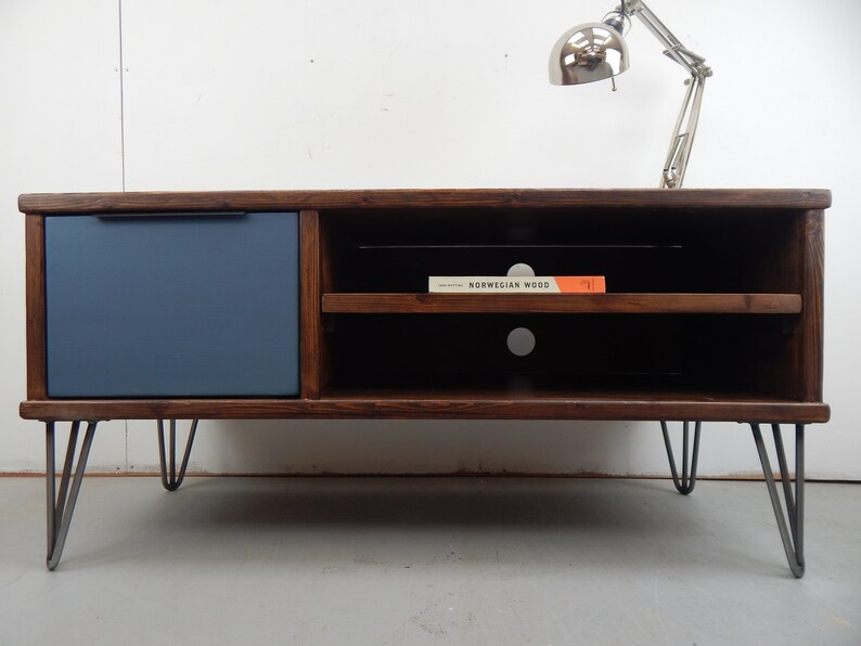 May include: A dark brown wooden TV stand with a blue painted drawer and two shelves. The stand has hairpin legs and is sitting on a light grey floor. A book titled "Norwegian Wood" is sitting on the top shelf.