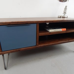 May include: A dark brown wooden TV stand with a blue drawer and two shelves. The stand has hairpin legs and a book titled "Norwegian Wood" on the top shelf.