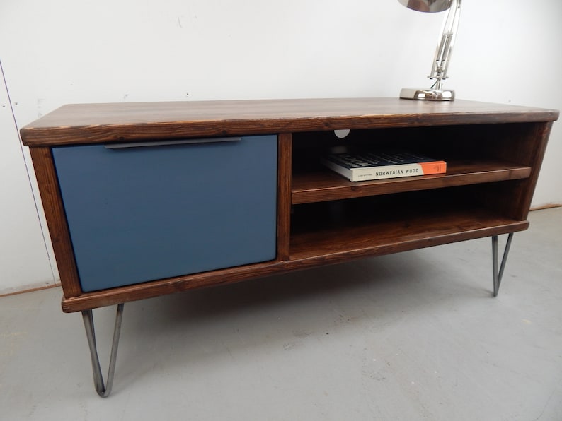 May include: A dark brown wooden TV stand with a blue drawer and two shelves. The stand has hairpin legs and a book titled "Norwegian Wood" on the top shelf.